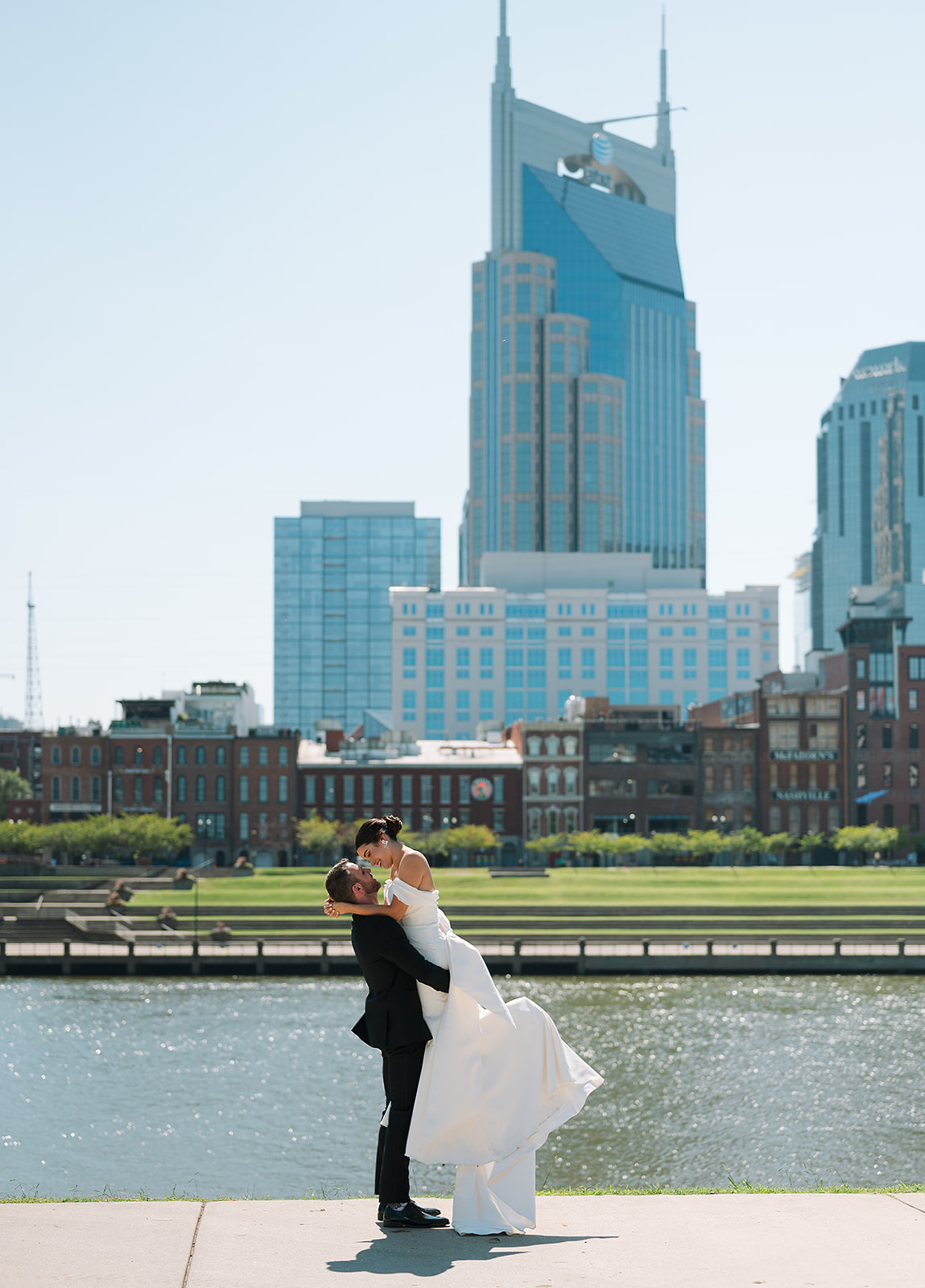 A groom lifts his new bride for a kiss while standing by cumberland river in downtown Nashville