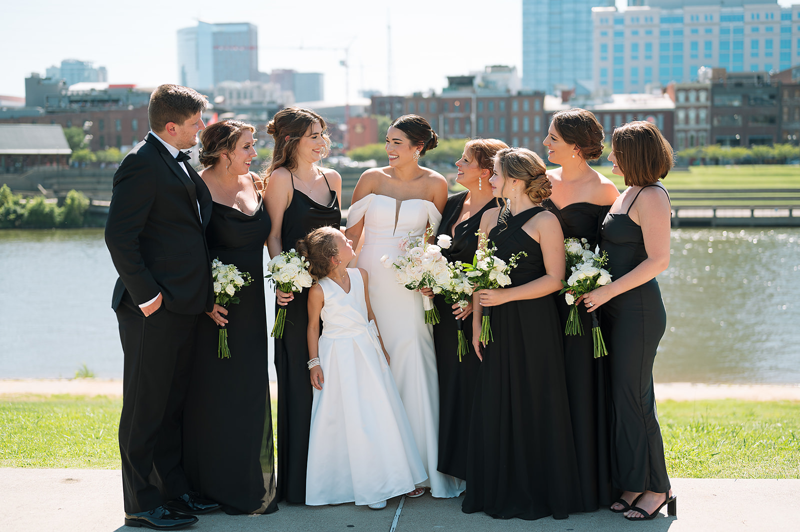 A bride stands with her bridesmaids and flower girl on the edge of a river