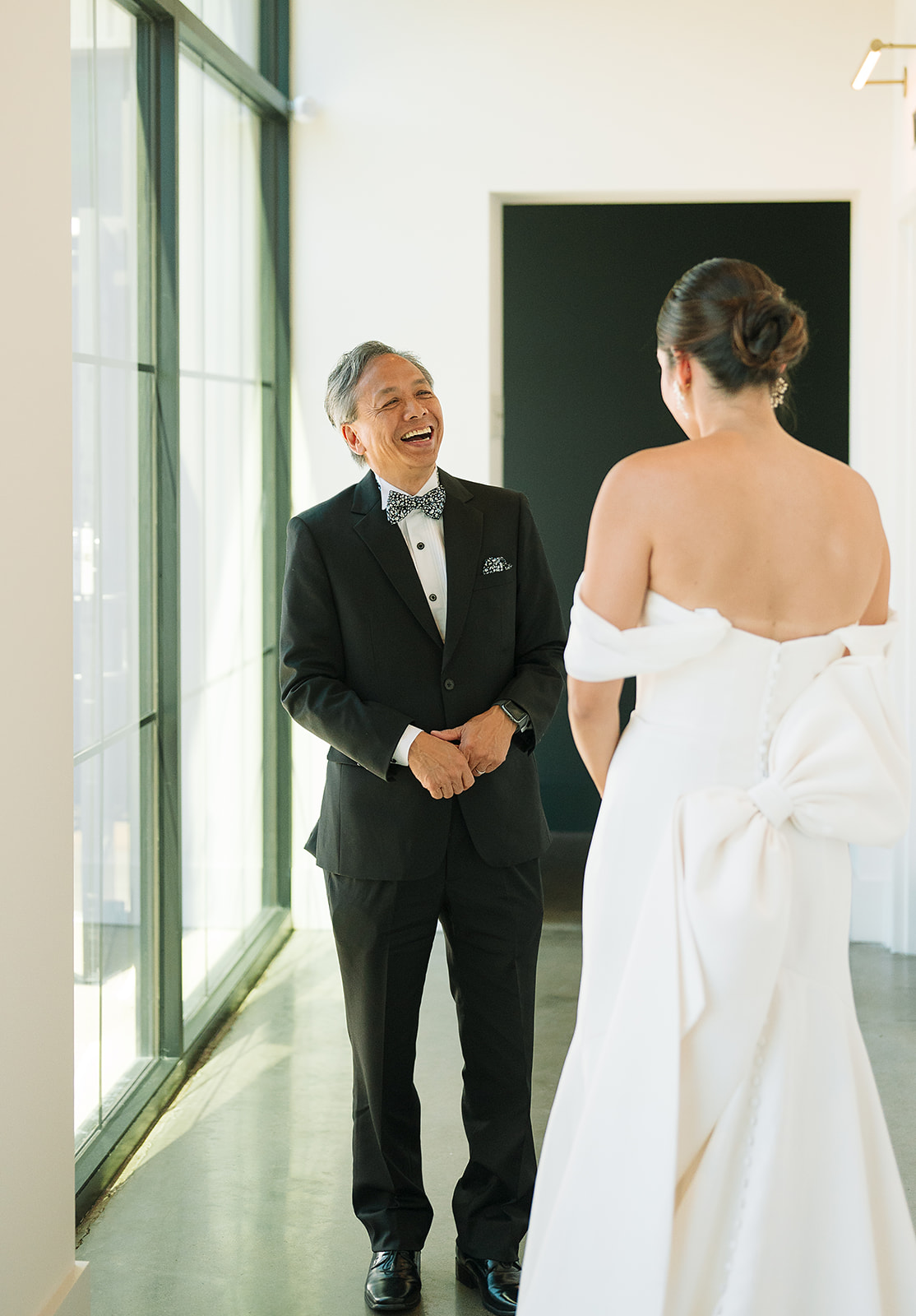 A father in a black suit laughs and smiles while during his first look with his daughter