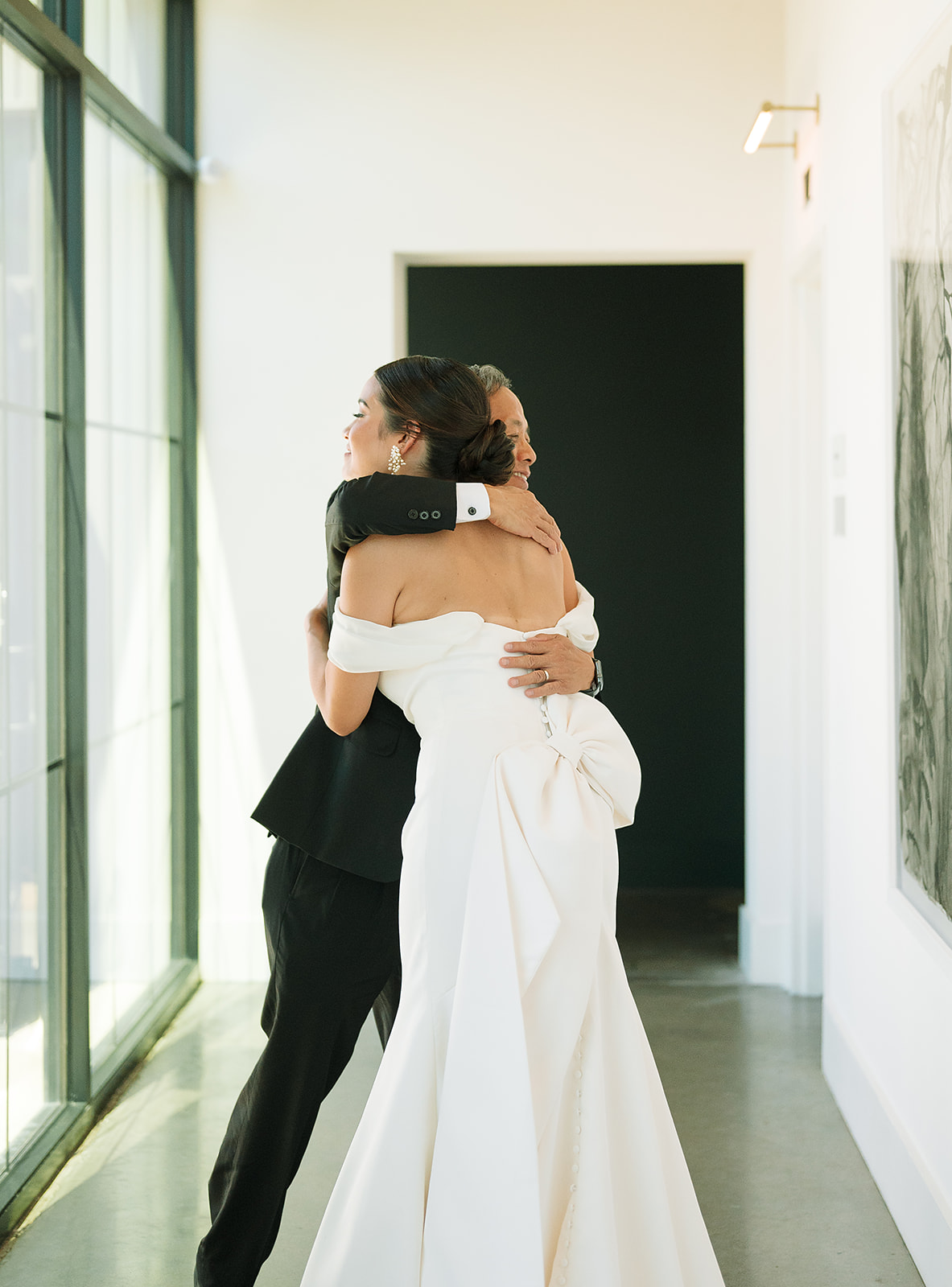 A father hugs his bride after seeing her in her dress for the first time in a hall by a large window