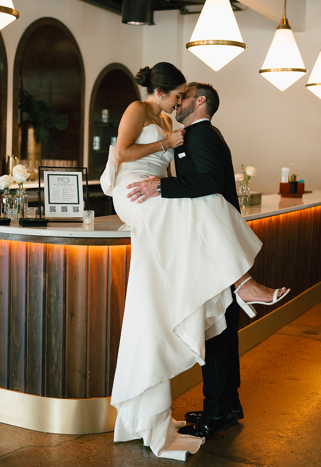 A bride sits on the bar while kissing her husband in a black suit at Saint Elle Wedding venue