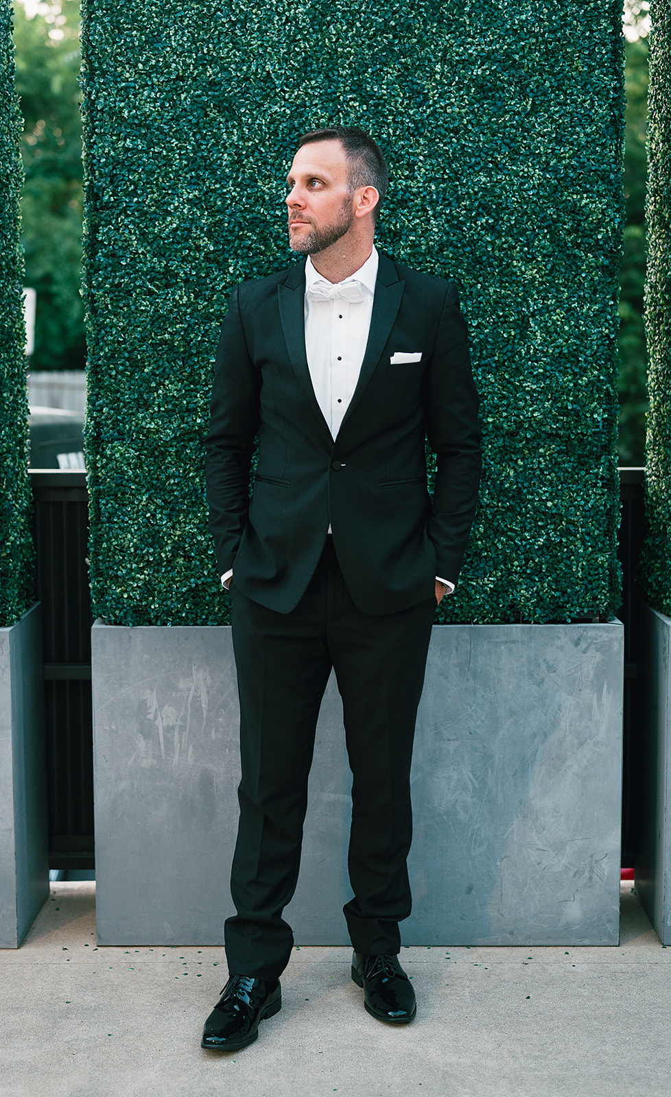 A groom stands with hands in pockets in a black suit on a patio with plant walls at a Saint Elle Wedding
